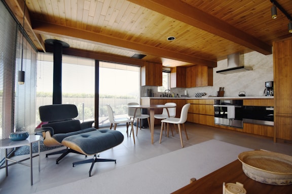 A modern kitchen and living area featuring wooden interior elements, an Eames lounge chair, a small round dining table with white chairs, stainless steel appliances, and large windows allowing natural light to brighten the space. The ceiling and cabinetry are made of wood, creating a warm and cozy atmosphere.