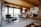 A modern kitchen and living area featuring wooden interior elements, an Eames lounge chair, a small round dining table with white chairs, stainless steel appliances, and large windows allowing natural light to brighten the space. The ceiling and cabinetry are made of wood, creating a warm and cozy atmosphere.