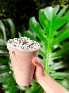 A hand holds a frothy cold beverage topped with whipped cream and chocolate sprinkles in a plastic cup. The background features large, vibrant green tropical leaves.