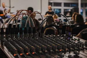 A mixing console with numerous knobs and wires is in the foreground, suggesting a live event or recording session. In the background, a blurred setting reveals people sitting and possibly watching a performance, with musical instruments barely visible.