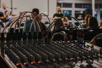 A mixing console with numerous knobs and wires is in the foreground, suggesting a live event or recording session. In the background, a blurred setting reveals people sitting and possibly watching a performance, with musical instruments barely visible.