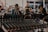 A mixing console with numerous knobs and wires is in the foreground, suggesting a live event or recording session. In the background, a blurred setting reveals people sitting and possibly watching a performance, with musical instruments barely visible.