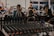 A mixing console with numerous knobs and wires is in the foreground, suggesting a live event or recording session. In the background, a blurred setting reveals people sitting and possibly watching a performance, with musical instruments barely visible.