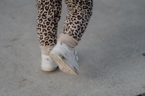 A pair of child's legs wearing leopard print leggings and white sneakers walking on a concrete surface.