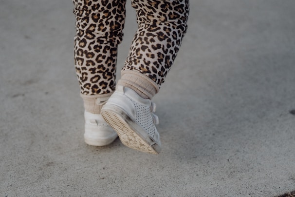 A pair of child's legs wearing leopard print leggings and white sneakers walking on a concrete surface.
