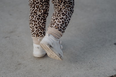A pair of child's legs wearing leopard print leggings and white sneakers walking on a concrete surface.