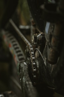 A close-up view of a bicycle chain and gear mechanism with a focus on the metal chain link and surrounding mechanical parts. The image highlights the details of the worn and slightly rusted components, illustrating the functional aspect of the bicycle's drivetrain.