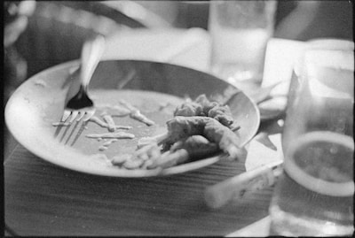 A black and white image depicting a partially eaten meal on a plate, with a fork resting on the edge. There are a few pieces of food left, and the scene includes a beverage glass and some blurred background elements, suggesting a dining setting.