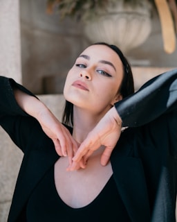 A person with dark hair and dramatic eye makeup poses with arms raised gracefully, creating a triangular shape under the chin. The background includes a blurred decorative element resembling an urn.