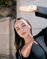 A young woman with long dark hair and striking eyeliner holds her hand up to shield her eyes from the sun. She is wearing a black top and blazer, standing against a stone wall under a bright light, creating shadows on her face.