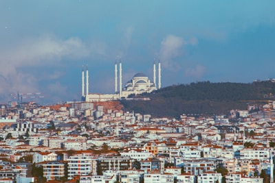 A large mosque with prominent minarets stands atop a hill, surrounded by dense urban development. The foreground is dominated by a sprawling cityscape of residential and commercial buildings. The background consists of a clear, blue sky with scattered clouds.