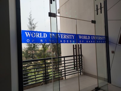 Glass doors with blue signage displaying 'World University of Bangladesh.' The background features a railing and some trees, indicating an outdoor view. The space appears to be part of a modern building design.