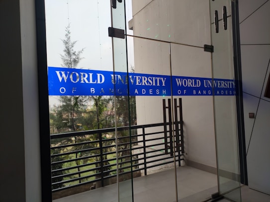 Glass doors with blue signage displaying 'World University of Bangladesh.' The background features a railing and some trees, indicating an outdoor view. The space appears to be part of a modern building design.