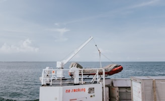 A calm ocean view with a ship's deck featuring a mounted crane, holding a covered inflatable boat. The ship's deck has a visible 'no smoking' sign in red, with a horizon line separating the sea from the clear sky.