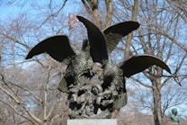 Two large bronze eagles with outstretched wings are perched on a stone pedestal, surrounded by leafless trees. An American flag can be seen in the background, slightly out of focus.