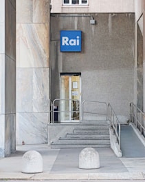 A modern entrance featuring a small blue sign with the word 'Rai' above a glass door. The area includes a short set of concrete steps and a ramp for accessibility, with metallic railings. The building exterior is a combination of marble and concrete textures, creating a simple and functional urban facade.