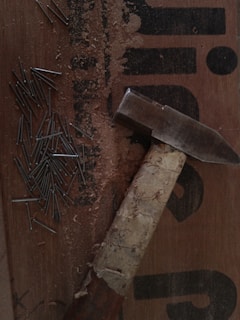 A collection of nails scattered on a wooden surface next to a hammer. The hammer has a well-used, wrapped handle and lies on a board with text partially visible. Wood shavings are also present, suggesting recent use.