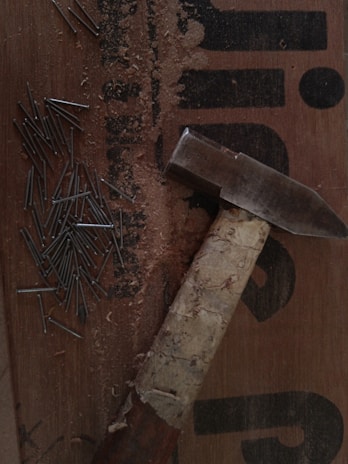 A collection of nails scattered on a wooden surface next to a hammer. The hammer has a well-used, wrapped handle and lies on a board with text partially visible. Wood shavings are also present, suggesting recent use.