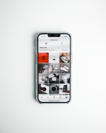 A smartphone displays a social media app with a grid of various post previews, showcasing different images and items such as shoes and accessories. The phone is placed on a clean, white surface.