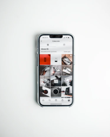 A smartphone displays a social media app with a grid of various post previews, showcasing different images and items such as shoes and accessories. The phone is placed on a clean, white surface.