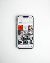 A smartphone displays a social media app with a grid of various post previews, showcasing different images and items such as shoes and accessories. The phone is placed on a clean, white surface.