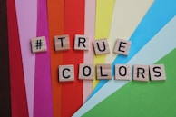 Scrabble tiles spelling out '#TRUE COLORS' are placed on a vibrant background of colored paper strips, including shades like red, pink, orange, yellow, blue, and green.