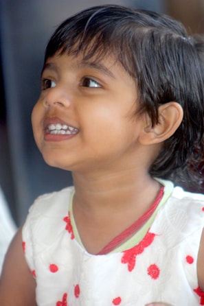 A young child is looking upwards with a cheerful expression, showcasing a wide smile. The child is dressed in a white outfit adorned with red flowers and dots, and has short dark hair.