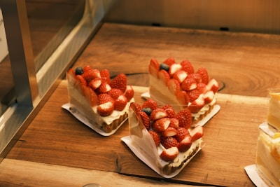 Three pieces of strawberry-topped cake sit on wooden trays. Each slice features numerous fresh strawberries and a couple of blueberries atop a creamy base, placed neatly on individual white paper liners.