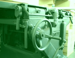 A close-up view of industrial machinery featuring a large wheel and several levers. The machinery has visible metallic components with an inscription on the side and is surrounded by stacked boxes.
