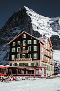 A picturesque mountain hotel with green shutters stands tall against a backdrop of a snowy, rugged mountain. The building is rustic and charming, with multiple floors and balconies. A group of people wearing winter gear sits outside on the snow, enjoying the sunny weather.