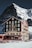A picturesque mountain hotel with green shutters stands tall against a backdrop of a snowy, rugged mountain. The building is rustic and charming, with multiple floors and balconies. A group of people wearing winter gear sits outside on the snow, enjoying the sunny weather.