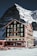 A picturesque mountain hotel with green shutters stands tall against a backdrop of a snowy, rugged mountain. The building is rustic and charming, with multiple floors and balconies. A group of people wearing winter gear sits outside on the snow, enjoying the sunny weather.