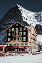 A picturesque mountain hotel with green shutters stands tall against a backdrop of a snowy, rugged mountain. The building is rustic and charming, with multiple floors and balconies. A group of people wearing winter gear sits outside on the snow, enjoying the sunny weather.