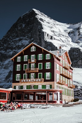 A picturesque mountain hotel with green shutters stands tall against a backdrop of a snowy, rugged mountain. The building is rustic and charming, with multiple floors and balconies. A group of people wearing winter gear sits outside on the snow, enjoying the sunny weather.