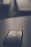 A smartphone is lying on a surface, displaying the Apple logo with a loading bar, suggesting it is in the process of booting up or restarting. The image is taken in low light with a blurred background, creating a minimalistic and focused composition.
