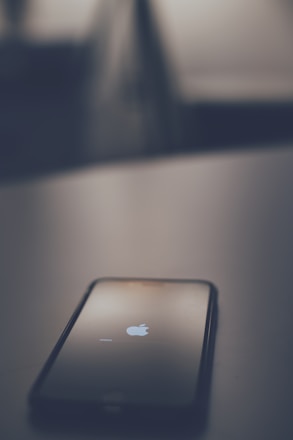 A smartphone is lying on a surface, displaying the Apple logo with a loading bar, suggesting it is in the process of booting up or restarting. The image is taken in low light with a blurred background, creating a minimalistic and focused composition.