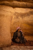 A man wearing traditional clothing, including a checkered headscarf, sits on a woven mat in a cave with sandstone walls. He is playing a stringed musical instrument and appears relaxed and contemplative.
