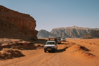 A desert landscape featuring rugged, rocky formations with three off-road vehicles traveling along a sandy path. The scene includes dry, arid terrain under a clear blue sky, with mountains in the background.