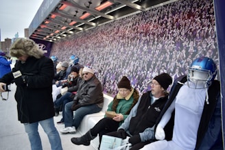 A group of people are seated on a bench under a shelter with heating elements above. They are dressed warmly, some wearing hats and gloves, indicating cold weather. A mannequin in football gear sits among them. The background features a large mural of a cheering crowd.