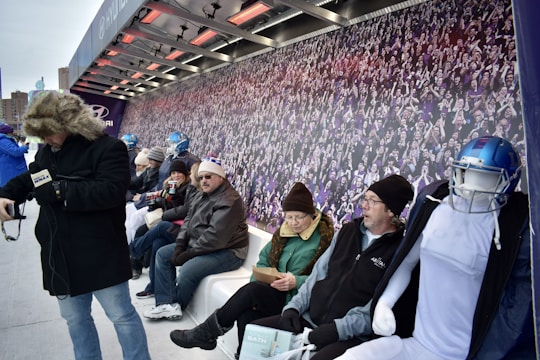 A group of people are seated on a bench under a shelter with heating elements above. They are dressed warmly, some wearing hats and gloves, indicating cold weather. A mannequin in football gear sits among them. The background features a large mural of a cheering crowd.