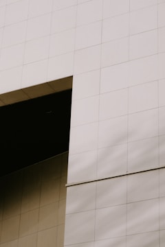 A close-up view of a modern building facade with a grid of large, smooth, pale beige tiles. The tiles are arranged in a geometric pattern, and there is a shadow running diagonally across them. A deep, dark opening is visible at the top left corner, adding contrast to the lighter surface.