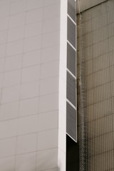 A modern architectural facade featuring large, white panels and vertical lines of dark vents. The building has a sleek, minimalist design with clean, geometric shapes. There is a netting or mesh structure on the adjacent side, giving a textured contrast to the smooth surface of the facade.