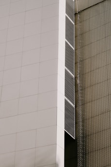 A modern architectural facade featuring large, white panels and vertical lines of dark vents. The building has a sleek, minimalist design with clean, geometric shapes. There is a netting or mesh structure on the adjacent side, giving a textured contrast to the smooth surface of the facade.