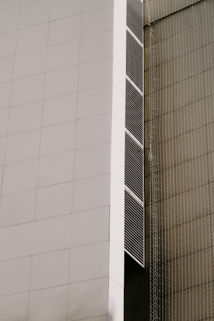 A modern architectural facade featuring large, white panels and vertical lines of dark vents. The building has a sleek, minimalist design with clean, geometric shapes. There is a netting or mesh structure on the adjacent side, giving a textured contrast to the smooth surface of the facade.