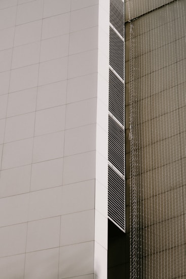 A modern architectural facade featuring large, white panels and vertical lines of dark vents. The building has a sleek, minimalist design with clean, geometric shapes. There is a netting or mesh structure on the adjacent side, giving a textured contrast to the smooth surface of the facade.