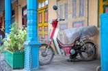 A vintage motorcycle is parked in front of a brightly colored, traditional building. The motorcycle is covered in a protective cloth with a wicker basket attached to its front. The backdrop features vibrant blue columns, colorful doors, and potted plants.
