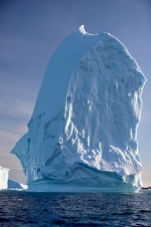 A massive iceberg rises from the ocean, exhibiting a rugged, towering shape with sharp edges and smooth surfaces. The iceberg's icy exterior glistens under sunlight, casting shadows and revealing a gradient of blues and whites. The calm water at its base reflects portions of the iceberg, adding to the serene atmosphere.