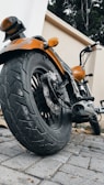 A close-up perspective of a motorcycle focusing on the rear tire and the bike's body. The orange paint of the motorcycle contrasts with the neutral tones of the cobblestone path and surrounding walls. The exhaust pipe and brake disc are visible, along with the handlebars and rearview mirror.
