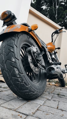 A close-up perspective of a motorcycle focusing on the rear tire and the bike's body. The orange paint of the motorcycle contrasts with the neutral tones of the cobblestone path and surrounding walls. The exhaust pipe and brake disc are visible, along with the handlebars and rearview mirror.