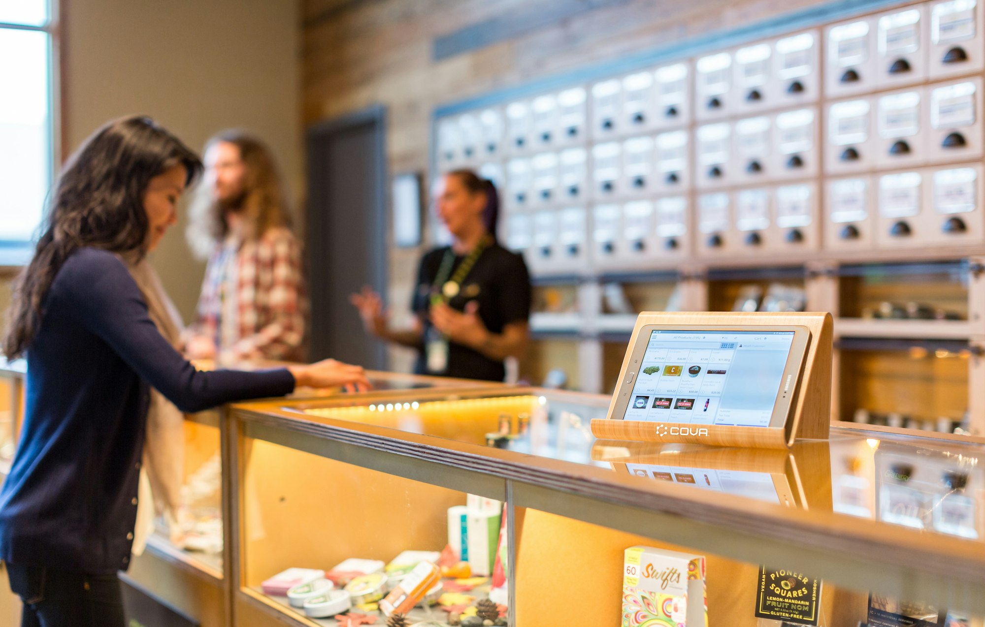 A lit-up screen displaying Cova's point of sale cannabis software is in focus atop a glass counter in a dispensary. In the background, a female cannabis customer peruses the options in the case while two budtenders help another consumer.